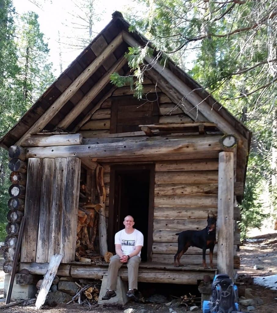 Man and his Doberman sitting on the front porch of a rustic cabin.